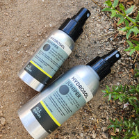 Two bottles of HYDROSOL  'JUNIPER'  lying on sandy ground next to green plants. The bottles are metallic silver with black spray nozzles and black/white labels with a yellow stripe.