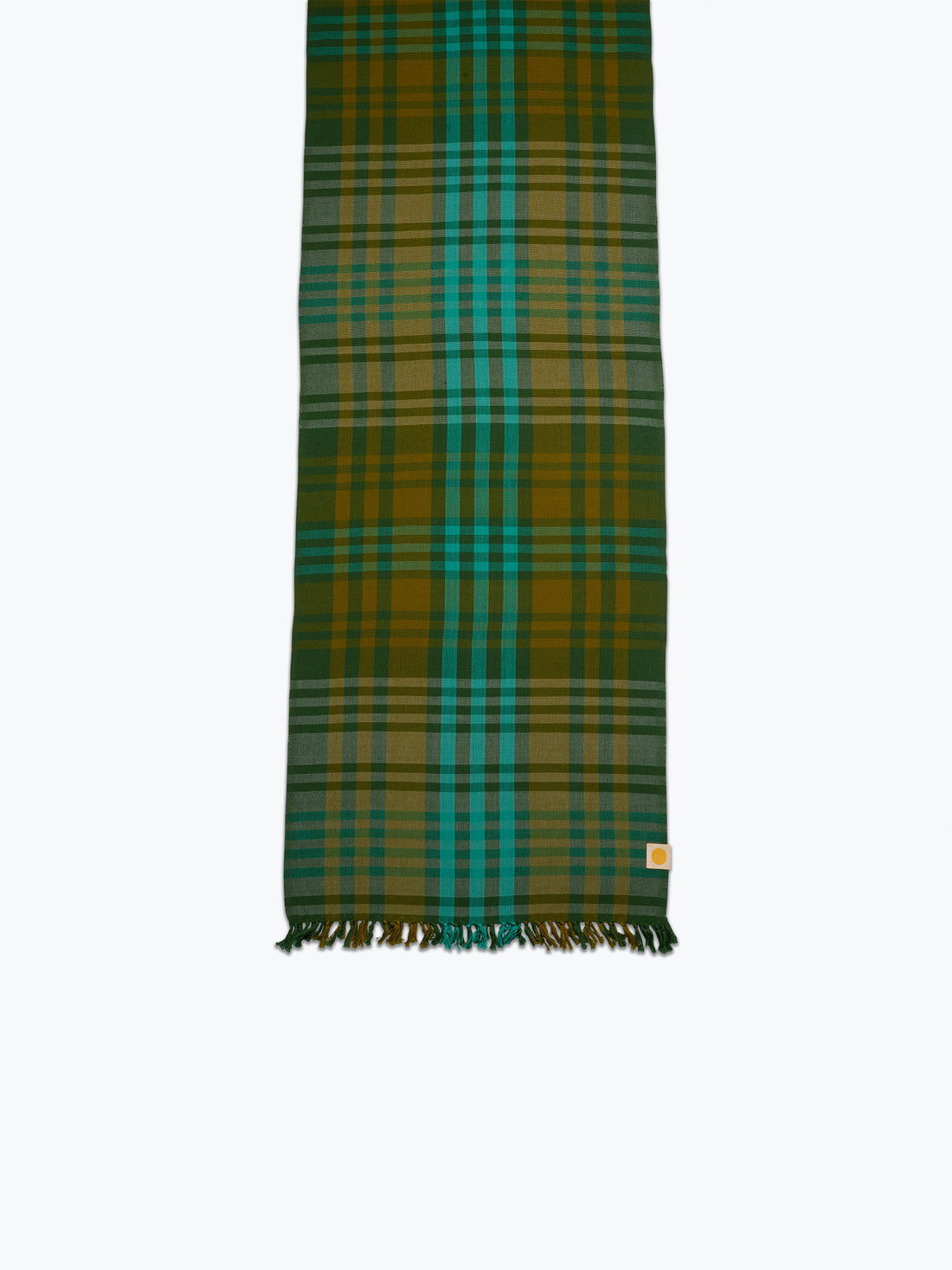 A long plaid tablelinen in green, teal, and mustard yellow with fringed ends, shown flat against a light gray background.