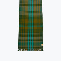 A long plaid tablelinen in green, teal, and mustard yellow with fringed ends, shown flat against a light gray background.