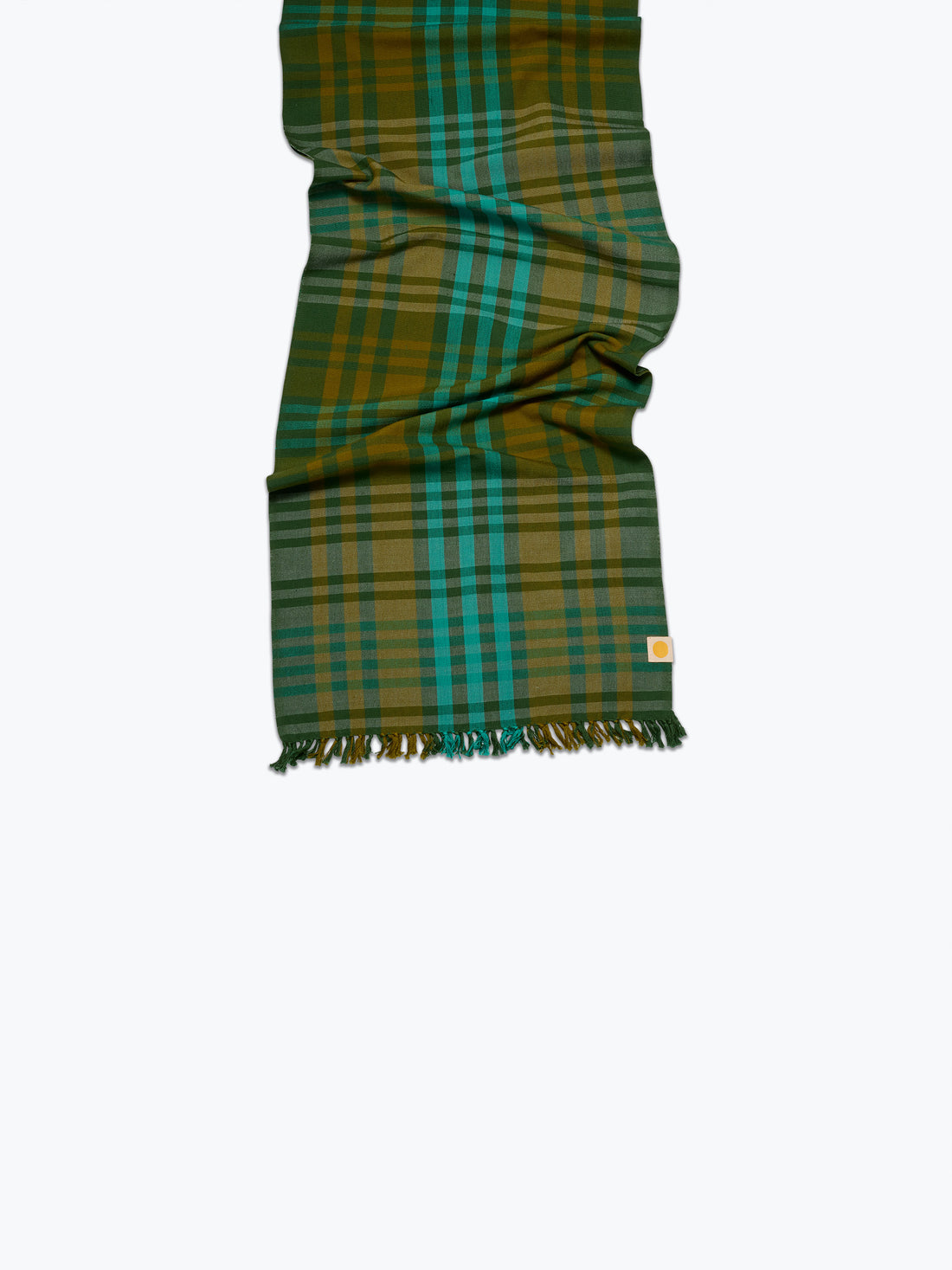 A folded plaid tablerunner in green, teal, and mustard yellow with fringed ends, shown against a plain white background.