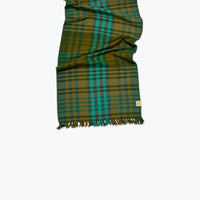 A folded plaid tablerunner in green, teal, and mustard yellow with fringed ends, shown against a plain white background.