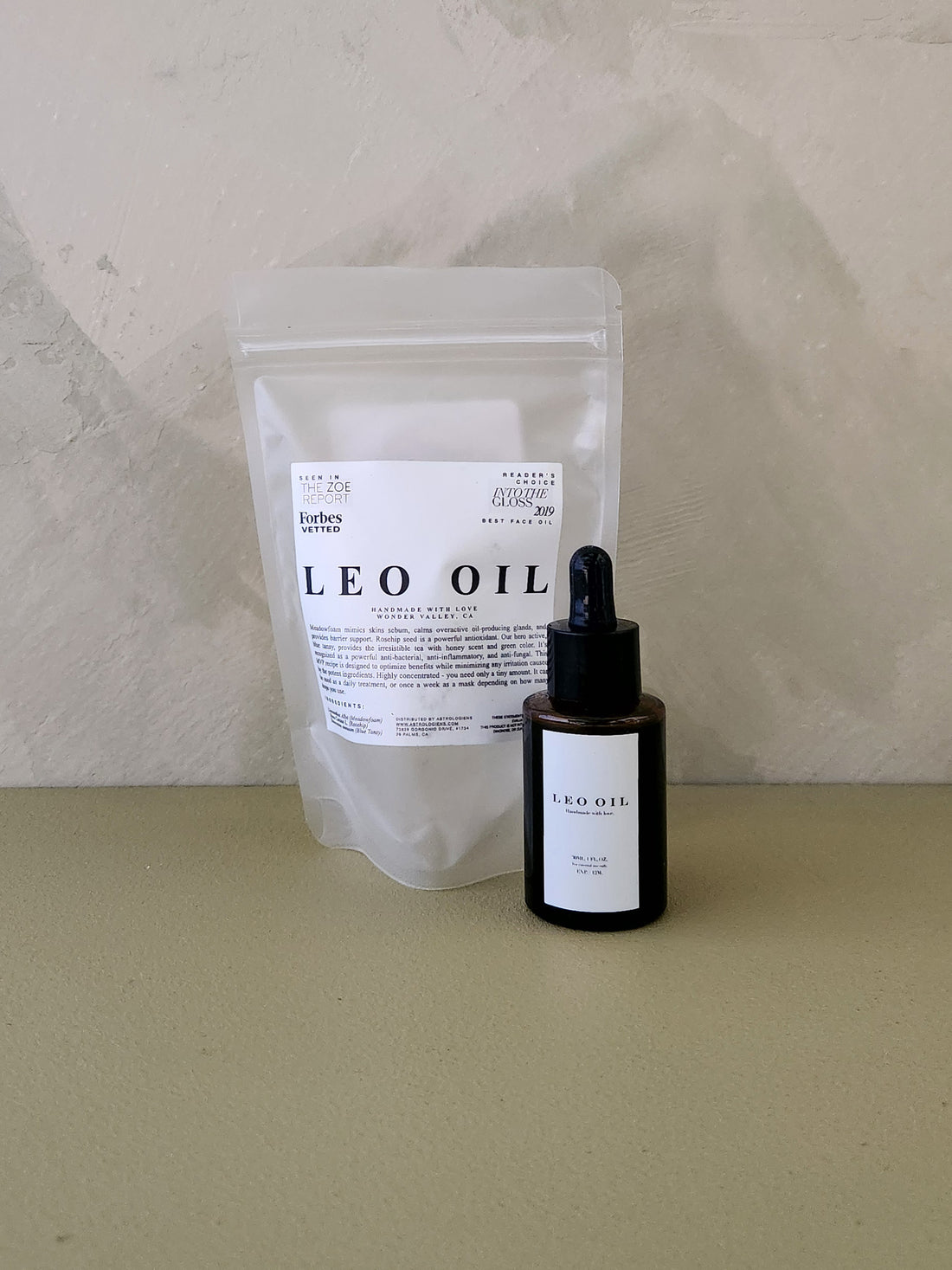 A small amber dropper bottle labeled LEO OIL stands in front of a translucent stand-up pouch with a white label that also reads LEO OIL, set on a beige surface against a textured beige wall.