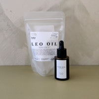A small amber dropper bottle labeled LEO OIL stands in front of a translucent stand-up pouch with a white label that also reads LEO OIL, set on a beige surface against a textured beige wall.