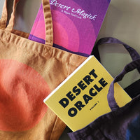 Two books titled 'Desert Magick' and 'Desert Oracle' with a brown tote bag on a gray background.