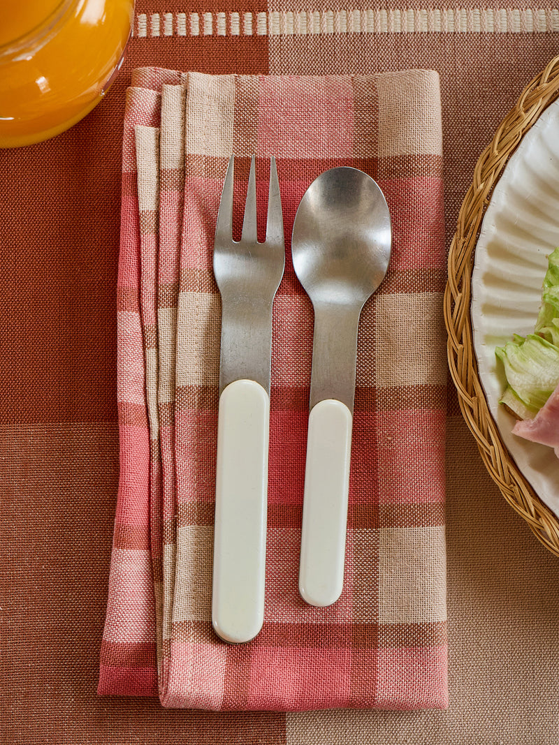 Silver fork and spoon on a pink and beige checkered napkin with a blurred plate of salad in the background.
