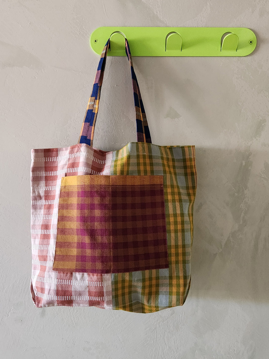 Plaid tote bag hanging on a green hook against a light gray wall.