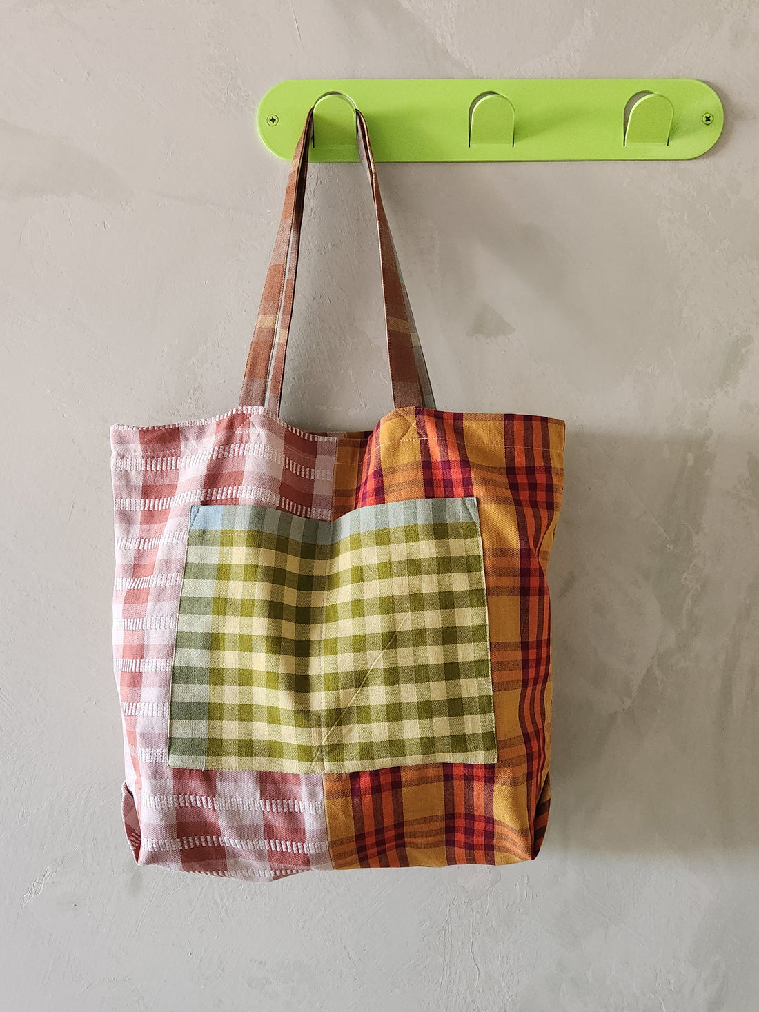 Colorful plaid tote bag hanging on a green hook against a plain wall.