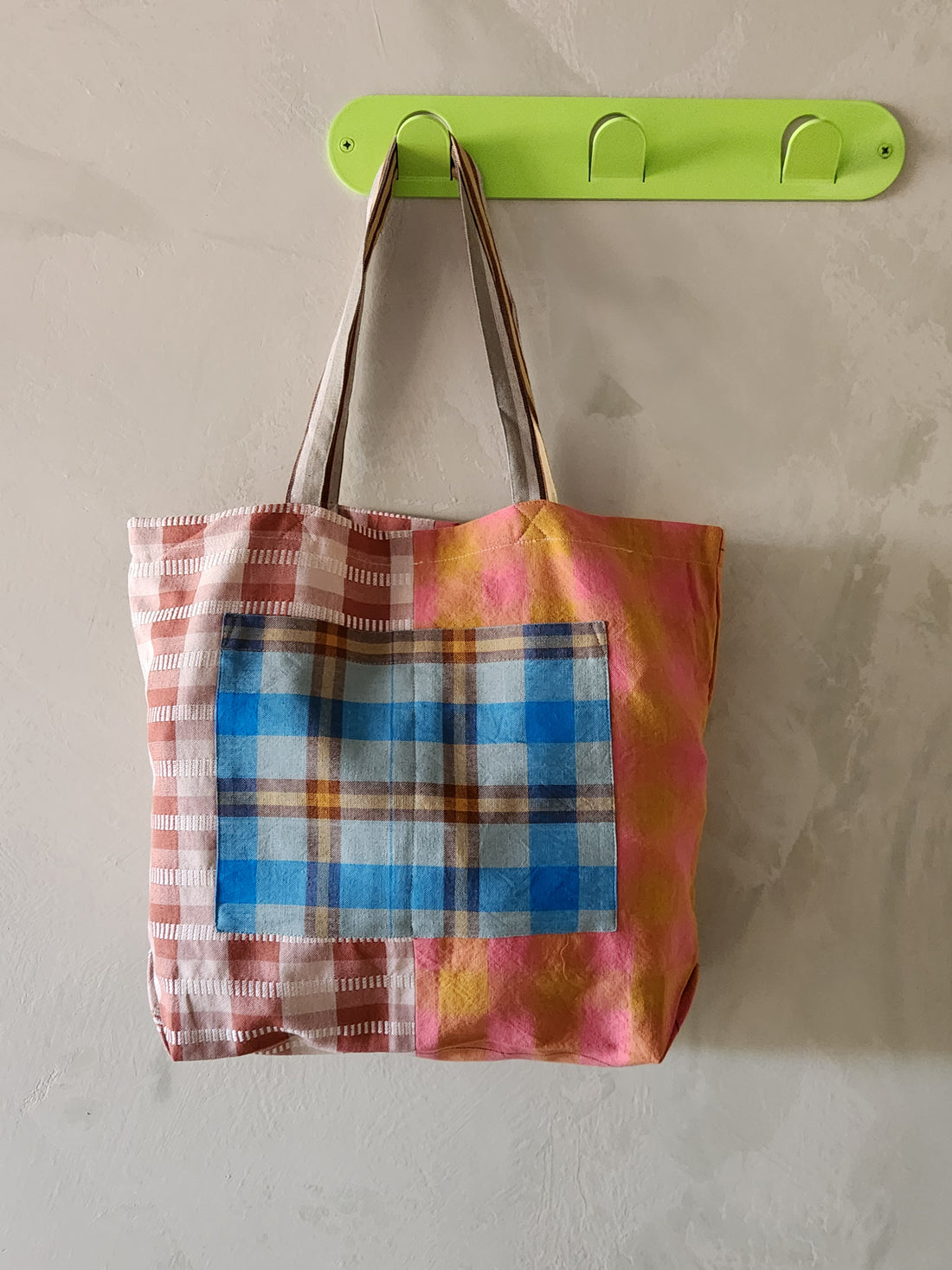 Colorful plaid tote bag hanging on a green hook against a beige wall.
