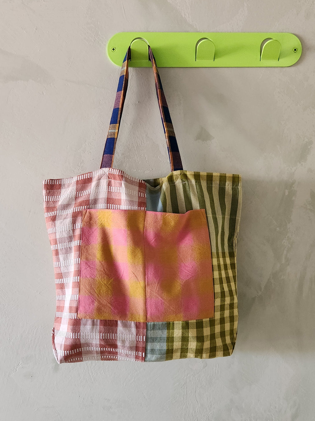 Colorful plaid tote bag hanging on a green wall hook against a plain background