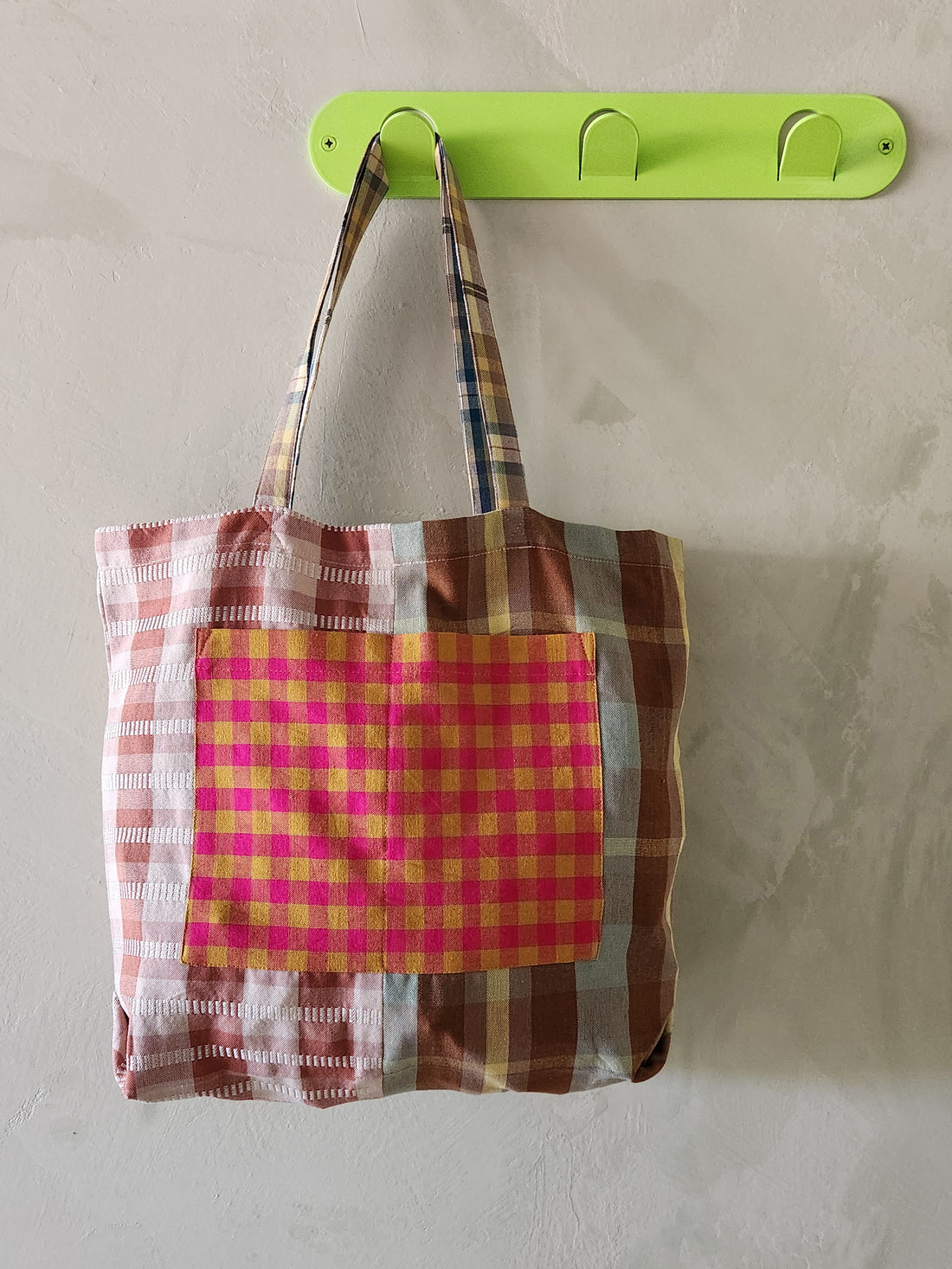 Plaid tote bag hanging on a green wall hook against a beige wall.