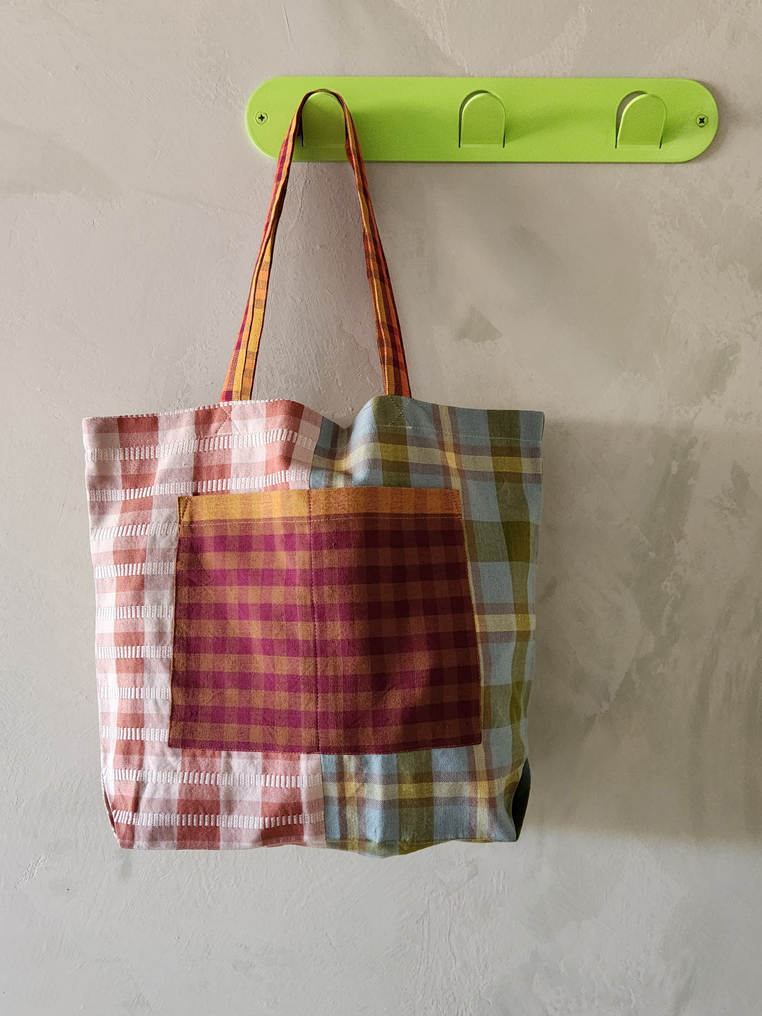 Plaid tote bag hanging on a green hook against a beige wall.