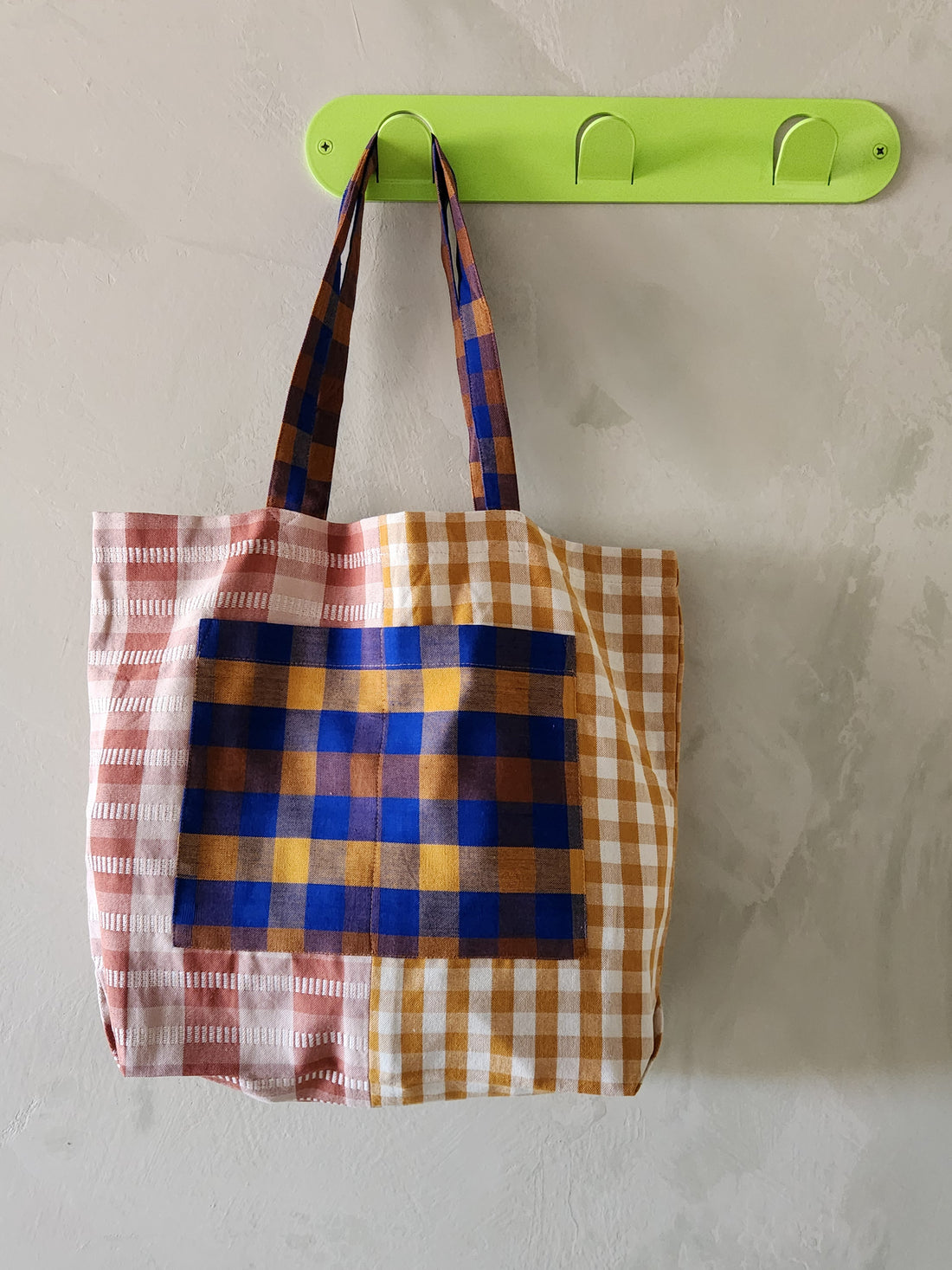 Plaid tote bag hanging on a green hook against a plain background