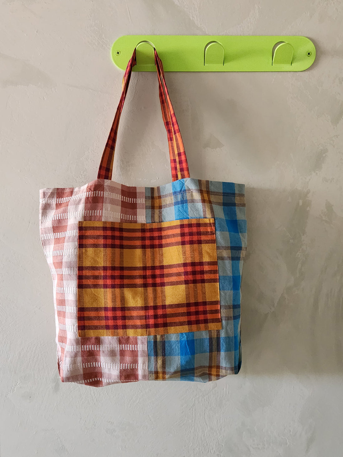 Plaid tote bag hanging on a green hook against a light gray wall.