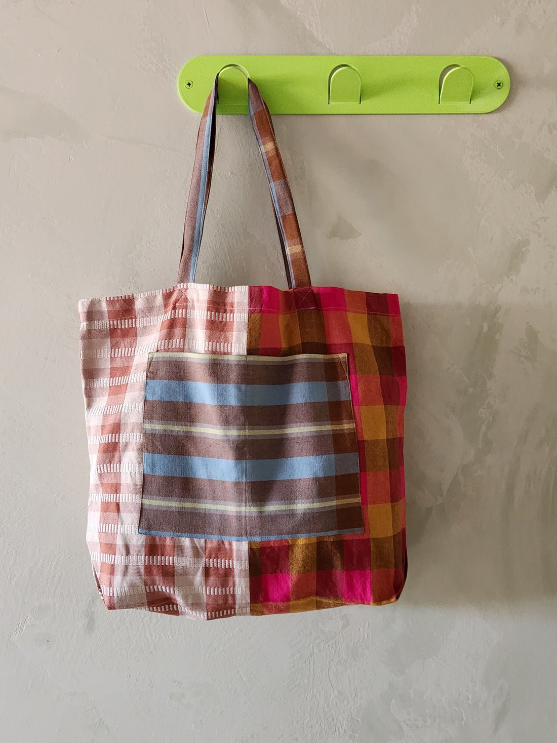 Colorful plaid tote bag hanging on a green wall hook against a plain background