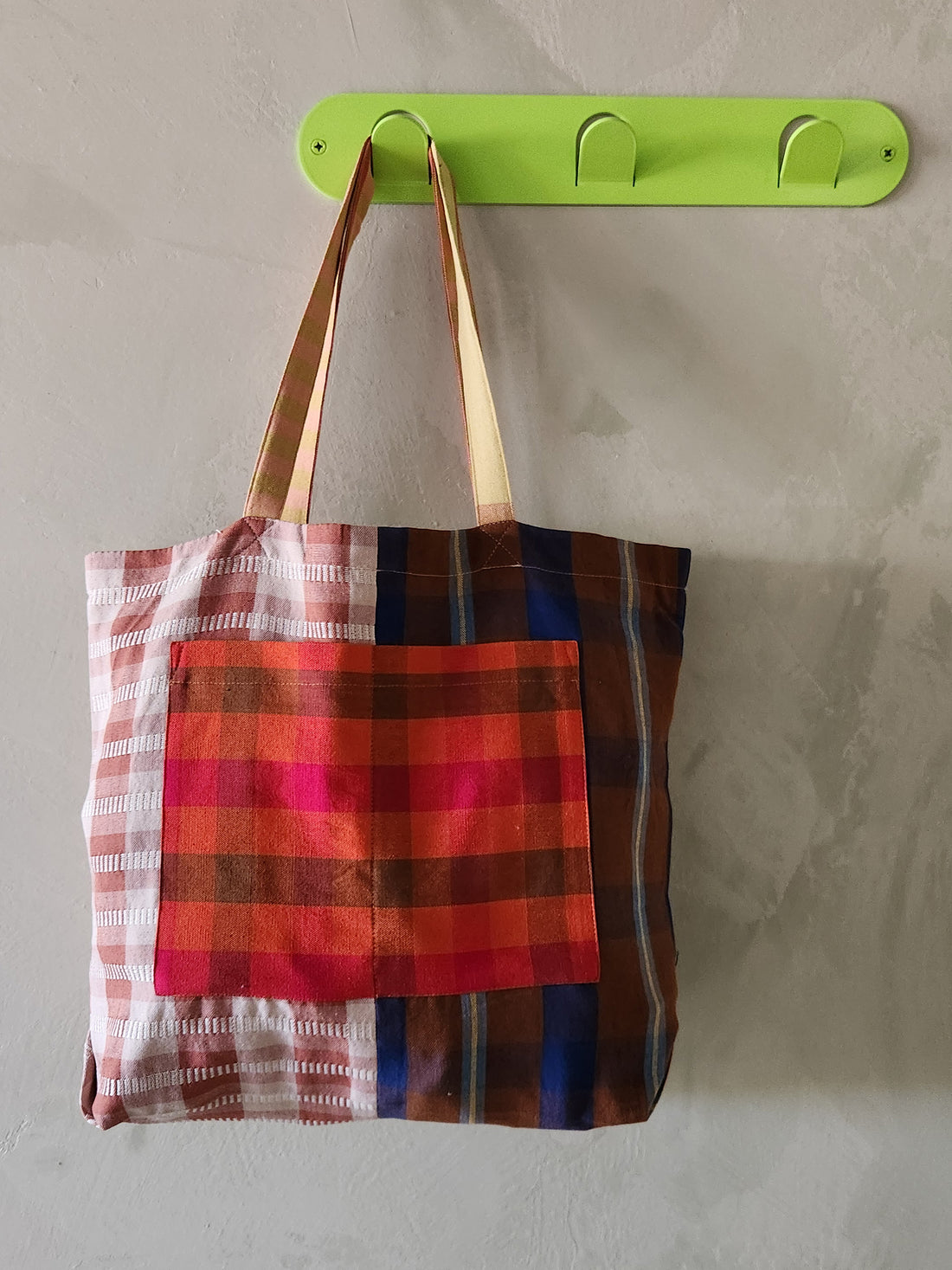 Colorful plaid tote bag hanging on a green wall hook against a neutral background