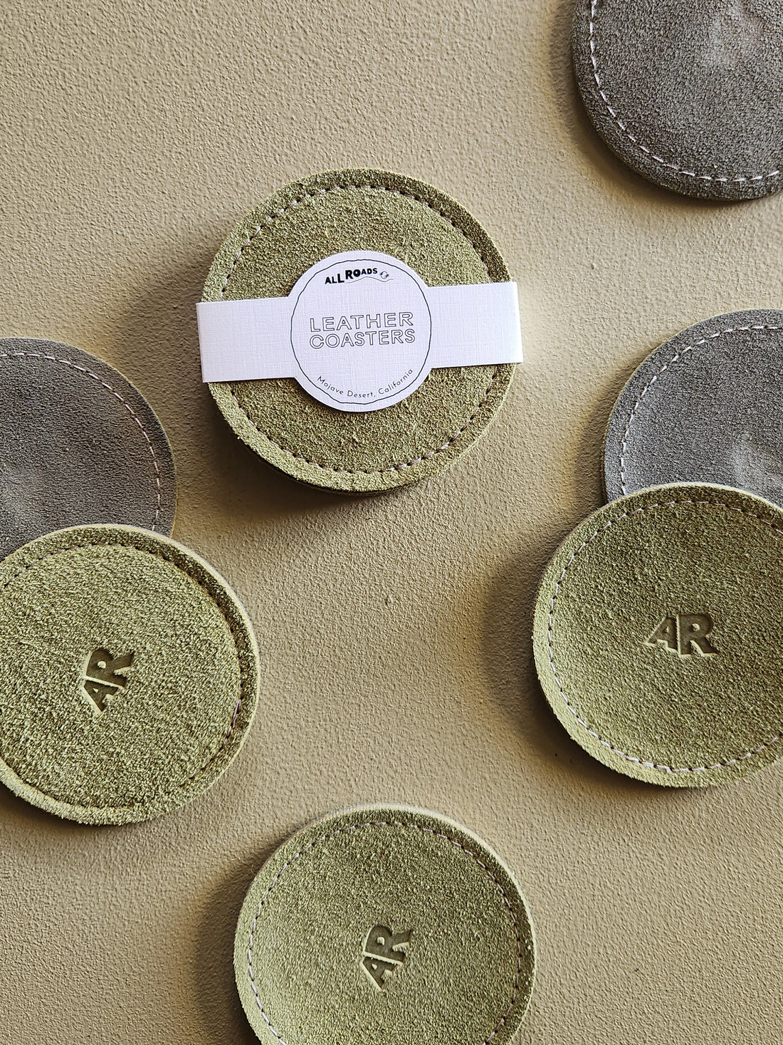 A set of round leather coasters arranged on a textured beige surface. One central coaster has a white paper band wrapped around it with a circular label that reads &