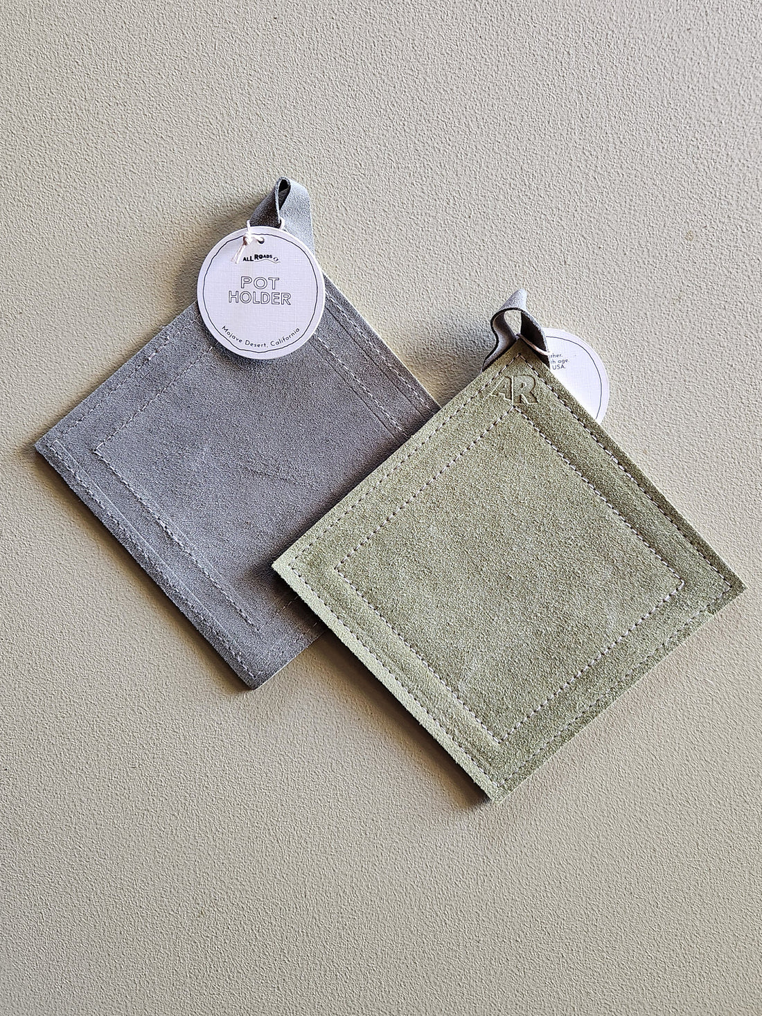 Two square fabric pot holders with stitched borders hang on a textured beige wall. One is gray and the other is light sage green, each with a white paper tag attached.