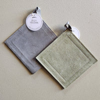 Two square fabric pot holders with stitched borders hang on a textured beige wall. One is gray and the other is light sage green, each with a white paper tag attached.