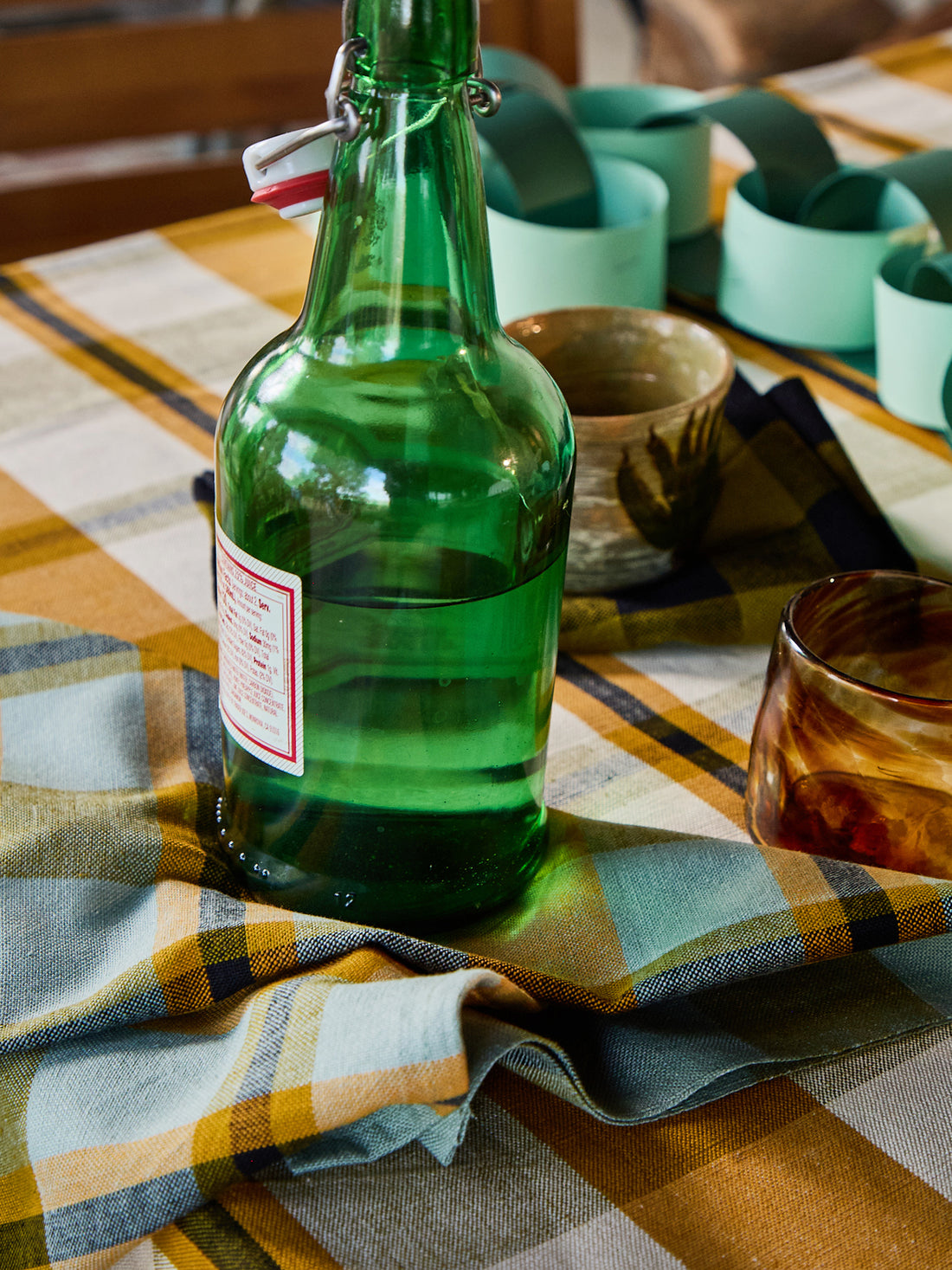 A green glass bottle with a swing-top closure and a white label, styled on a plaid tablecloth with matching green and amber glassware.