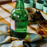 A green glass bottle with a swing-top closure and a white label, styled on a plaid tablecloth with matching green and amber glassware.