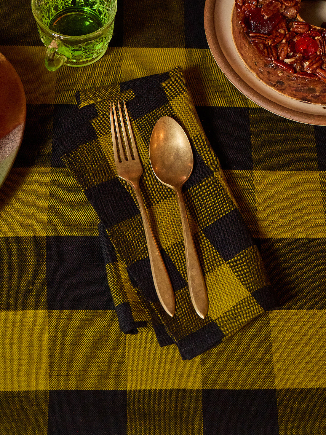 A rustic tabletop setting featuring a folded buffalo plaid napkin in olive green and black, styled with gold-toned metal cutlery (a fork and spoon) on top. The background shows a matching plaid tablecloth, a glass of green beverage, and a plate with food.