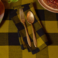 A rustic tabletop setting featuring a folded buffalo plaid napkin in olive green and black, styled with gold-toned metal cutlery (a fork and spoon) on top. The background shows a matching plaid tablecloth, a glass of green beverage, and a plate with food.