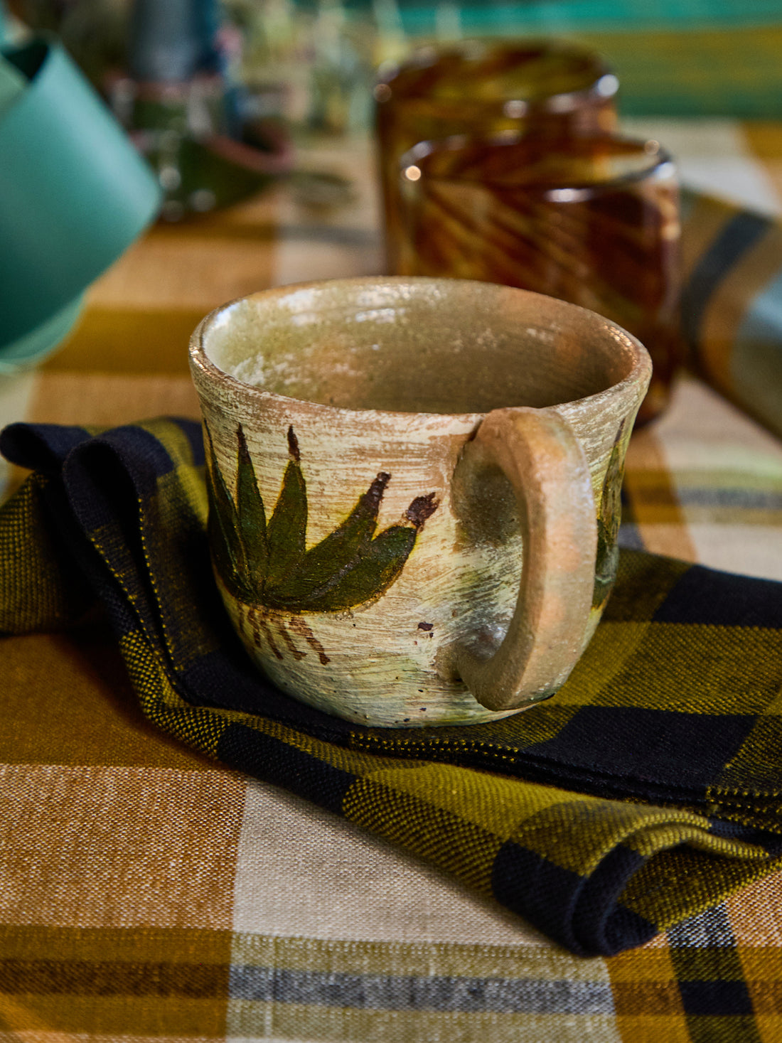 A rustic handmade ceramic mug with a green leaf design, resting on a black and olive green plaid napkin over a plaid tablecloth.