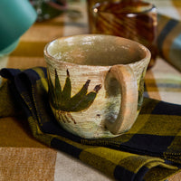 A rustic handmade ceramic mug with a green leaf design, resting on a black and olive green plaid napkin over a plaid tablecloth.