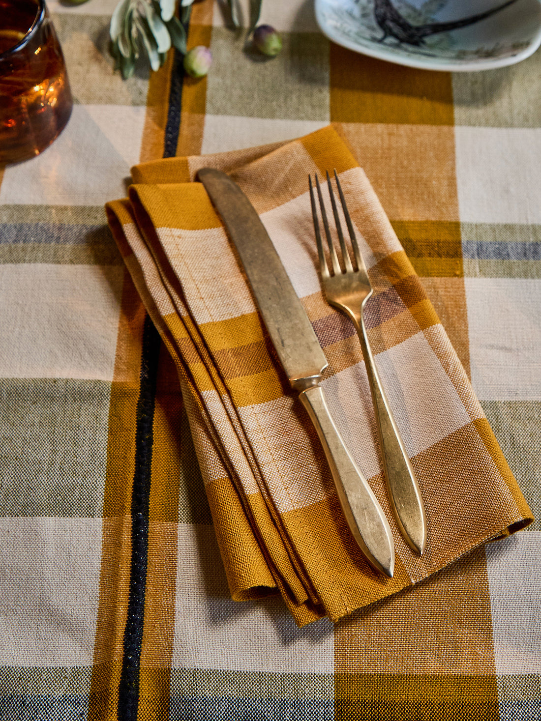 A folded linen napkin in a mustard yellow, beige, and brown plaid pattern, styled with a vintage-style knife and fork on a matching plaid tablecloth.