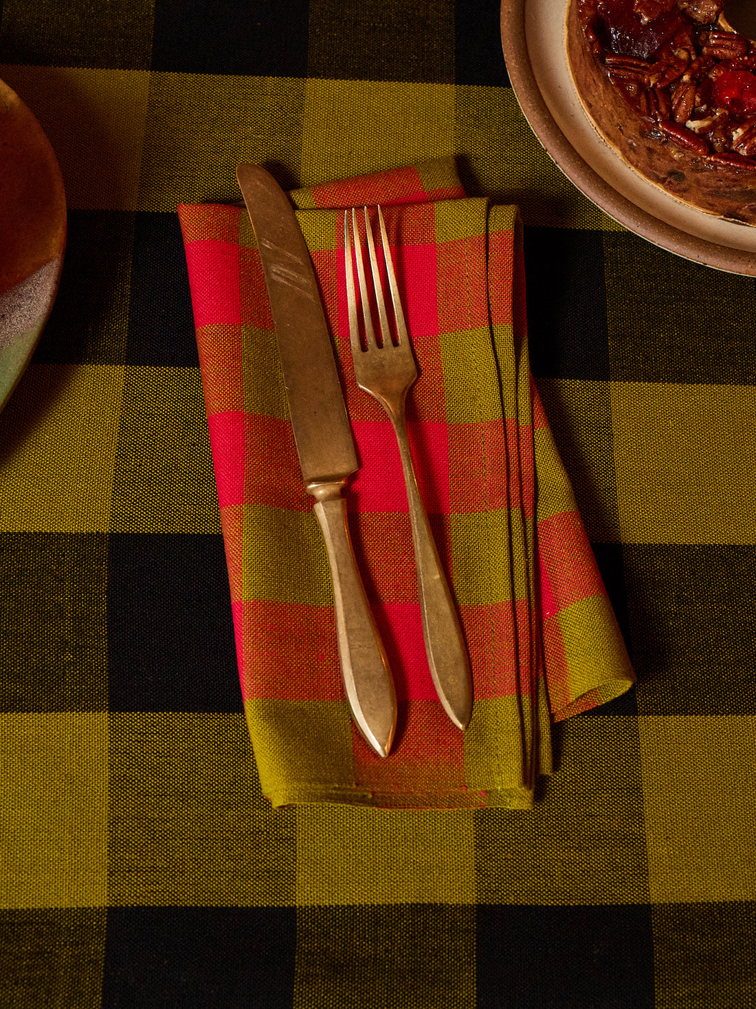 A folded red and olive green plaid napkin with a vintage-style gold knife and fork, set on a matching plaid tablecloth.