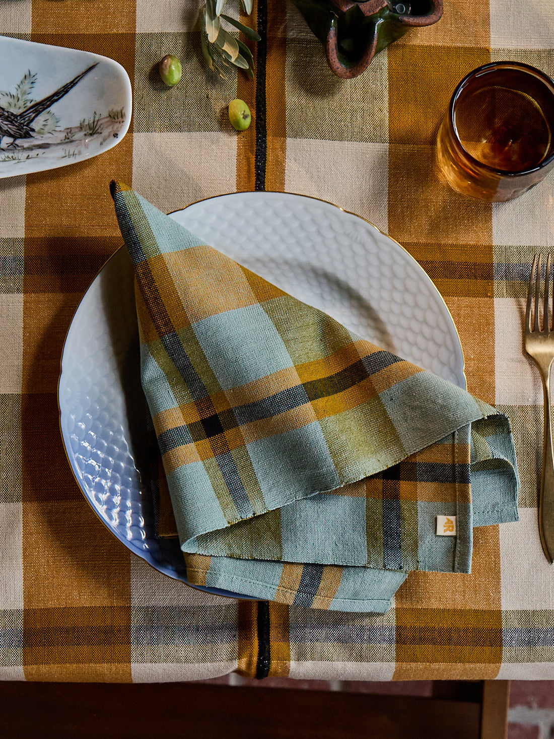 A folded plaid napkin in muted sage green, mustard yellow, and navy is arranged on a white plate at a table setting with a matching plaid tablecloth, gold-toned cutlery, and a small decorative dish.