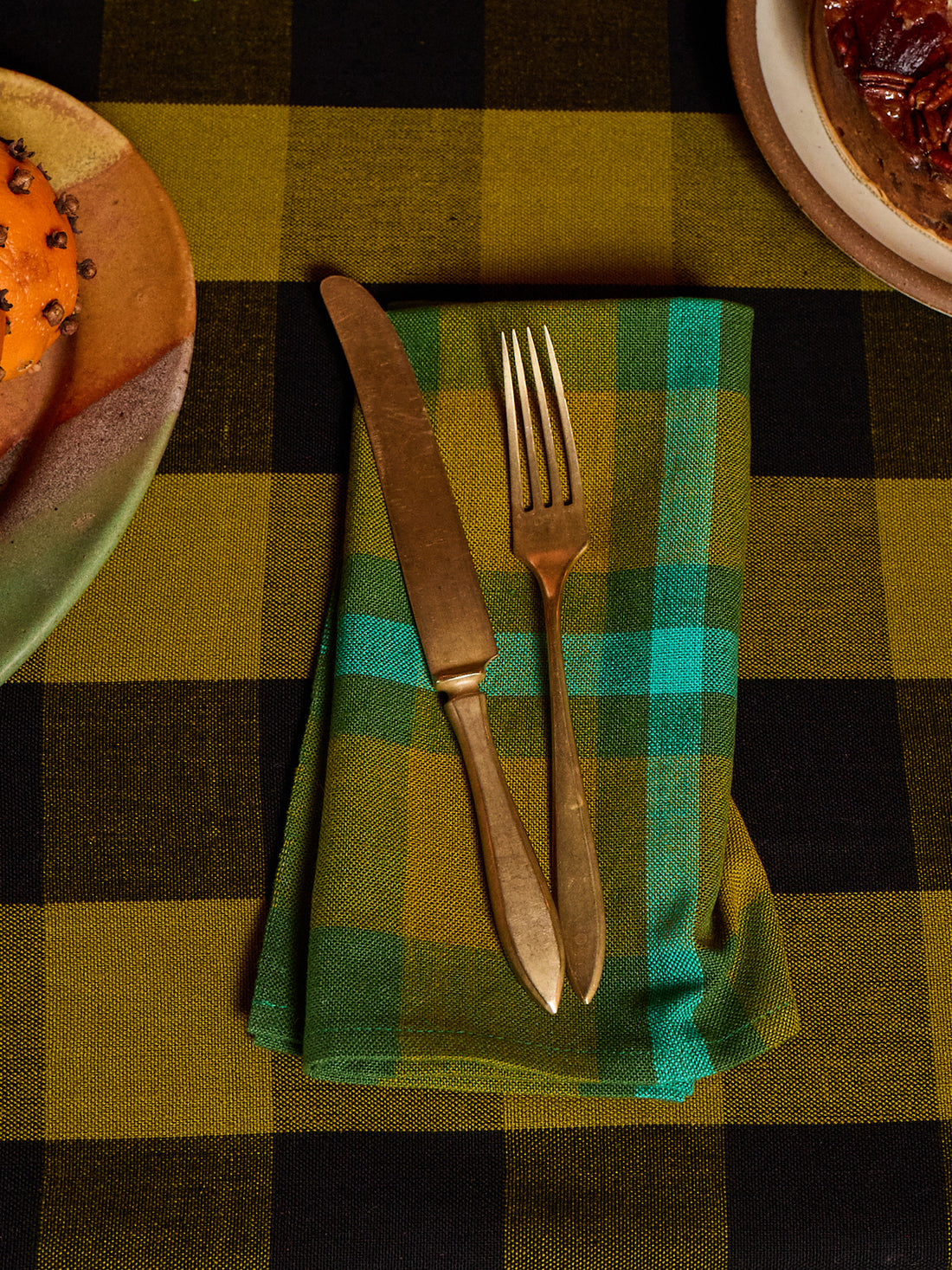 A folded green and teal plaid napkin with a vintage-style gold-toned knife and fork, set on a matching green and black buffalo plaid tablecloth.