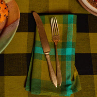 A folded green and teal plaid napkin with a vintage-style gold-toned knife and fork, set on a matching green and black buffalo plaid tablecloth.