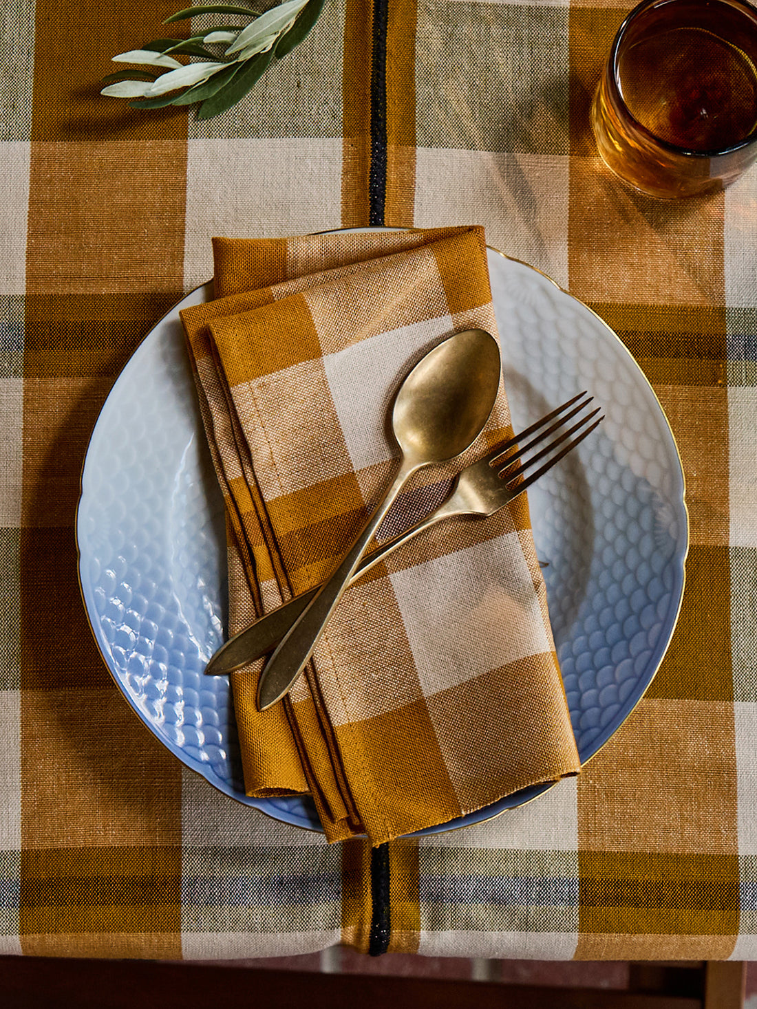A styled tabletop setting featuring a folded mustard yellow and cream plaid napkin on a white textured plate, with a gold spoon and fork resting on top, all set on a matching plaid tablecloth.