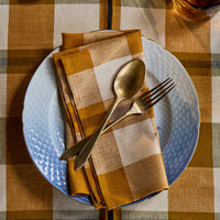 A styled tabletop setting featuring a folded mustard yellow and cream plaid napkin on a white textured plate, with a gold spoon and fork resting on top, all set on a matching plaid tablecloth.