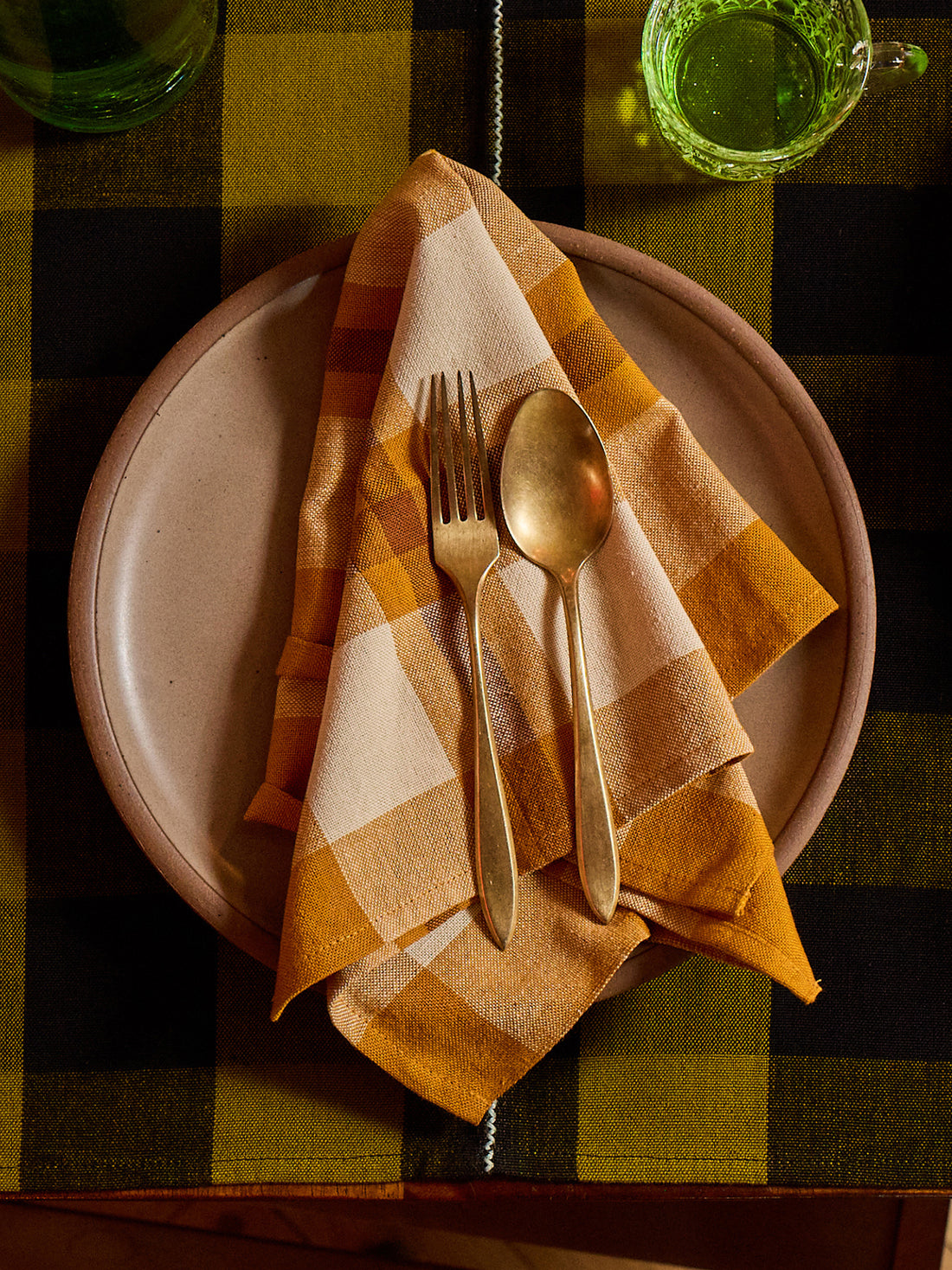 A styled tabletop setting featuring a folded ochre yellow and cream plaid napkin on a beige ceramic plate, with gold-toned cutlery. The setting is placed on a dark green and black buffalo plaid tablecloth, with a green glass mug visible in the background.