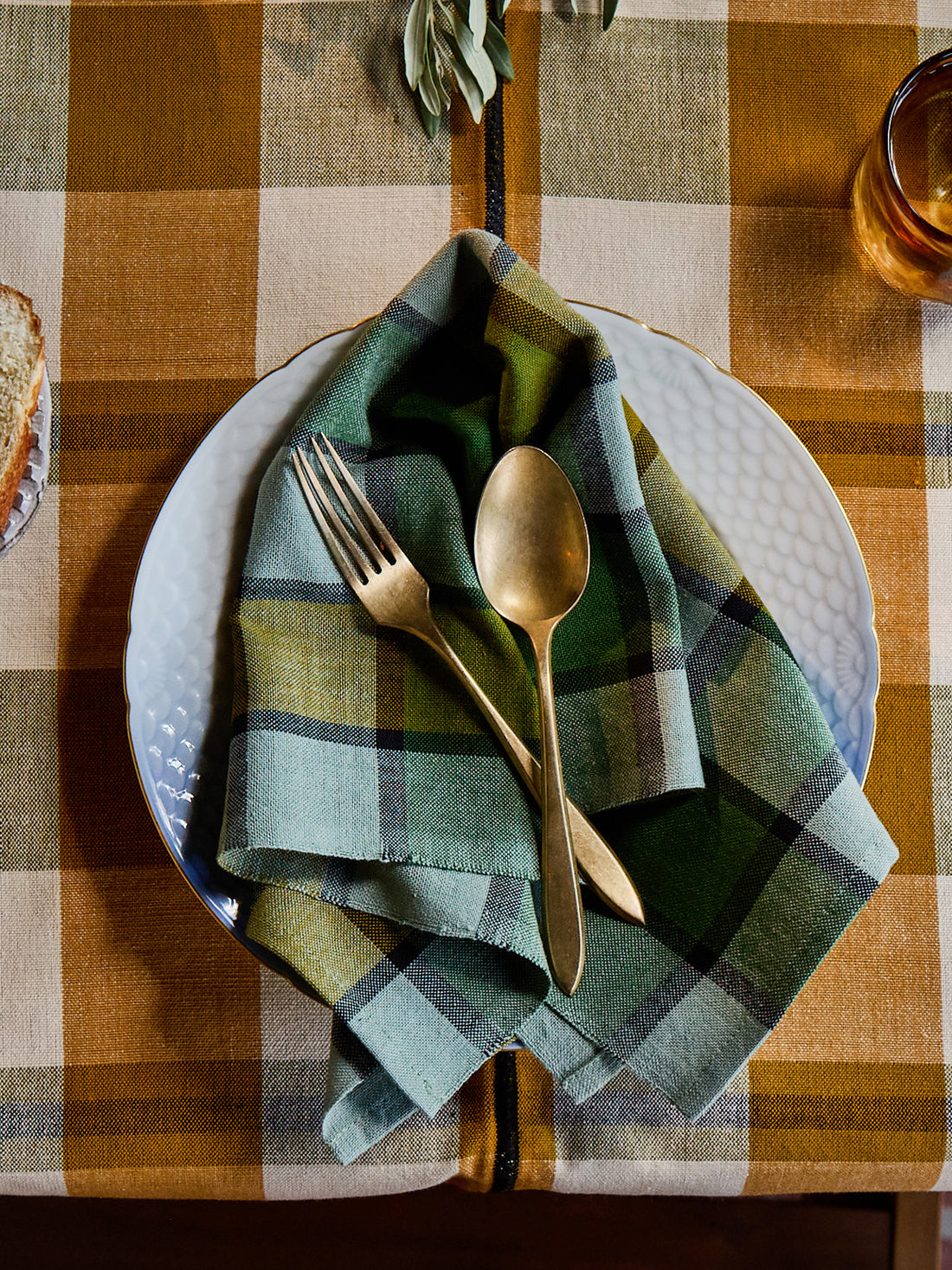 A folded plaid napkin in muted green, blue, and beige tones, layered over a white textured plate with gold-toned cutlery, set on a rustic brown and beige checkered tablecloth.