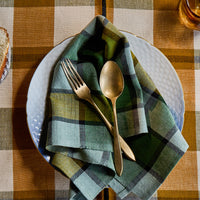 A folded plaid napkin in muted green, blue, and beige tones, layered over a white textured plate with gold-toned cutlery, set on a rustic brown and beige checkered tablecloth.
