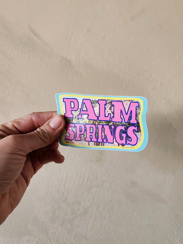 Hand holding a 'Palm Springs' sticker against a beige background
