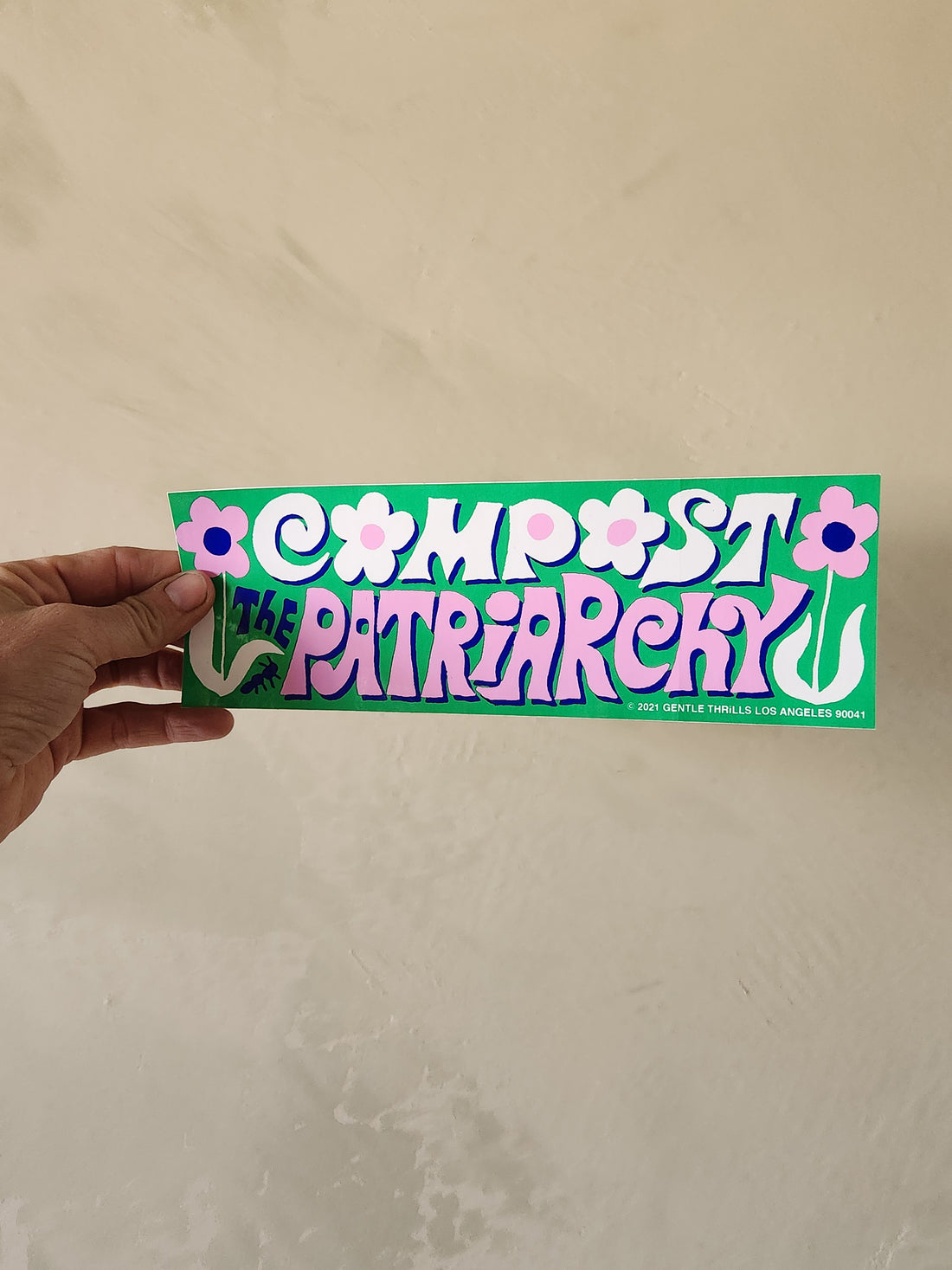 Hand holding a bumper sticker with text and floral designs on a beige background