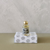A small glass perfume bottle with a gold cap and a black-and-white label reading 'FERAL MILK'  displayed on top of a white box with black brain illustrations against a textured light gray wall and beige surface.