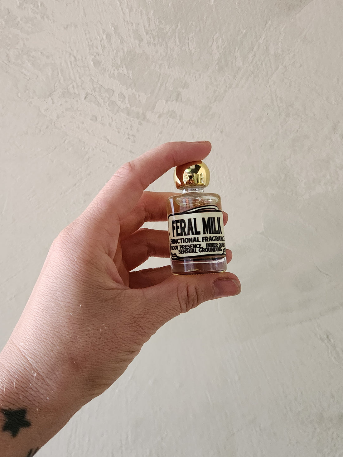 A hand holding a small glass bottle of Feral Milk Functional Fragrance with a gold cap against a textured off-white wall.