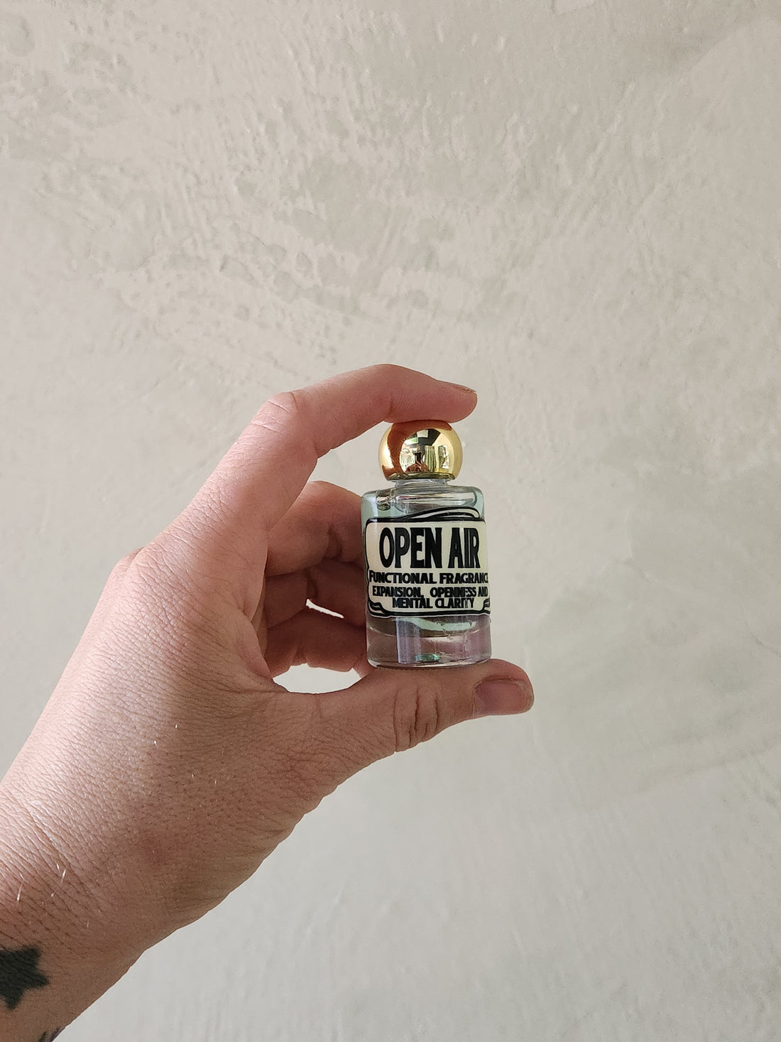 A hand holding a miniature glass bottle of perfume with a gold cap and a label that reads &