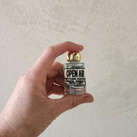 A hand holding a miniature glass bottle of perfume with a gold cap and a label that reads 'OPEN AIR'. The background is a textured off-white wall.