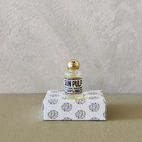 A small glass bottle with a gold cap and a black-and-white label reading 'SUN PULP' sits on top of a white box printed with black brain-like illustrations. The scene is set against a textured light gray wall and a beige surface.