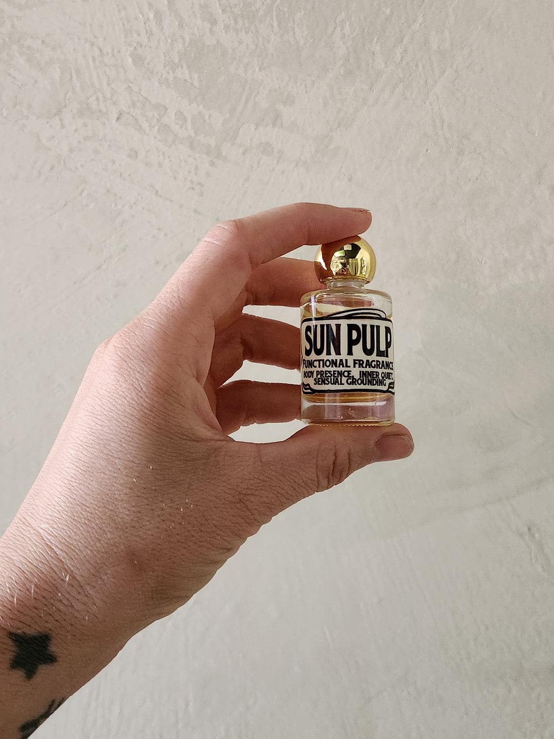 A hand holding a miniature glass bottle of Sun Pulp Functional Fragrance with a gold cap against a textured off-white wall.