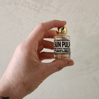A hand holding a miniature glass bottle of Sun Pulp Functional Fragrance with a gold cap against a textured off-white wall.