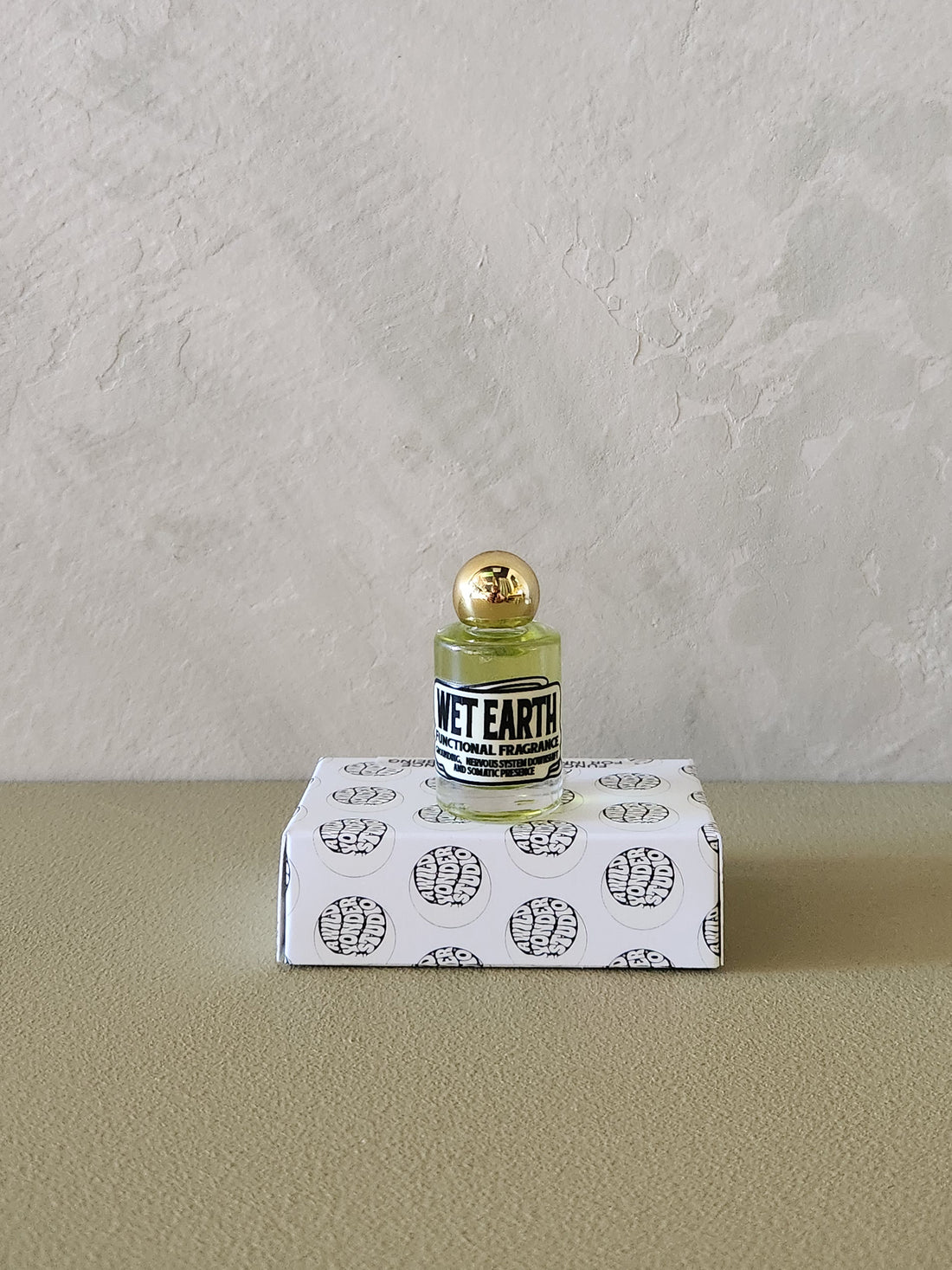 A small clear glass bottle with pale yellow liquid and a gold cap sits on top of a white box printed with black brain illustrations. The bottle label reads &