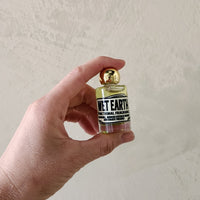 A hand holding a small glass bottle of Wet Earth Functional Fragrance with a gold cap against a textured off-white wall.