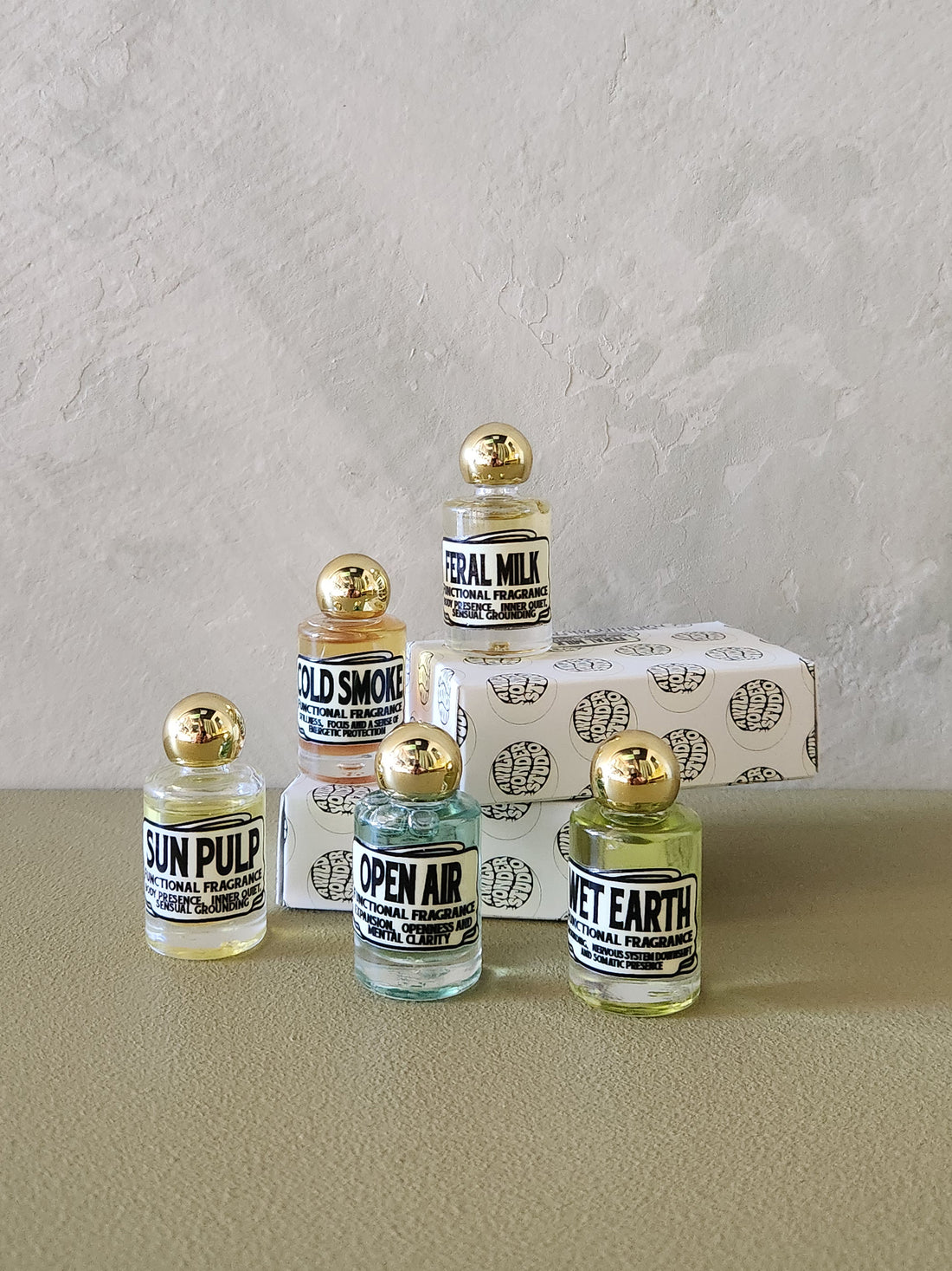 Five small glass perfume bottles with gold caps and black-and-white labels are arranged in front of a textured light gray wall. The labels read Sun Pulp, Cold Smoke, Neural Mix, Open Air, and Wet Earth.